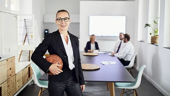 [Translate to Englisch:] Christiane Pütz hält Basketball und lächelt, im Hintergrund findet ein Meeting statt.