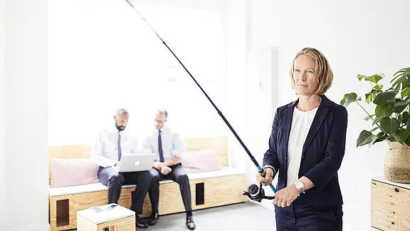 [Translate to Englisch:] Anja Wenzel mit "fischt" mit einer Angel im Büro, im Hintergrund eine Besprechung.