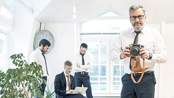 [Translate to Englisch:] Dirk Pütz hält eine Fotokamera hoch, im Hintergrund eine Besprechung.